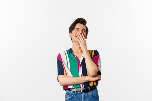 Image of handsome young man laughing over joke, having fun, cover mouth with hands and chuckle, standing over white background.