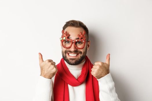 Christmas, New Year and celebration concept. Happy and satisfied man with beard, wearing party glasses, showing thumbs up in approval or like, standing over white background.
