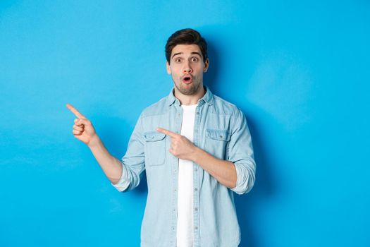 Portrait of surprised adult man in casual clothes showing announcement, pointing fingers left and looking amazed, standing against blue background.