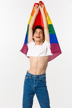 Vertical view of proud and happy gay man raising lgbtq rainbow flag, smiling with relieved emotion, wearing crop top with jeans, white background.