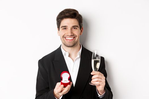 Close-up of handsome man in suit, making a proposal, giving engagement ring and raising glass of champagne, standing against white background.