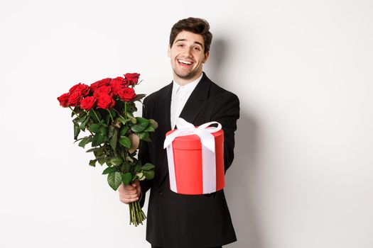 Concept of holidays, relationship and celebration. Image of handsome smiling guy in black suit, holding bouquet of red roses and giving you a gift, standing against white background.