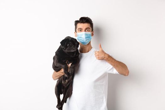 Covid-19, animals and quarantine concept. Young man in medical mask holding cute black pug dog, showing thumb up, like and approve, standing over white background.