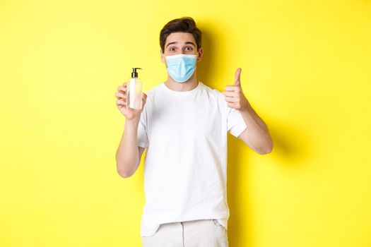 Concept of covid-19, quarantine and lifestyle. Satisfied young man in medical mask showing good hand sanitizer, thumbs up and recommending antiseptic, yellow background.