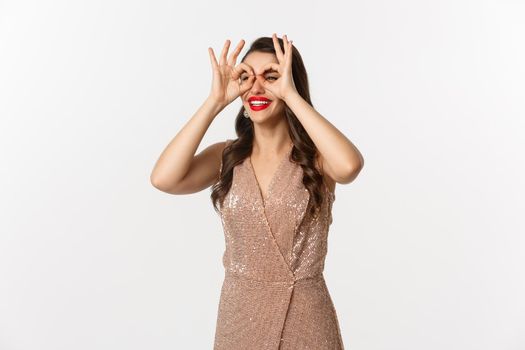 Christmas party and celebration concept. Young beautiful woman smiling happy, seeing something on distance, looking through hand binoculars, wearing elegant dress, white background.