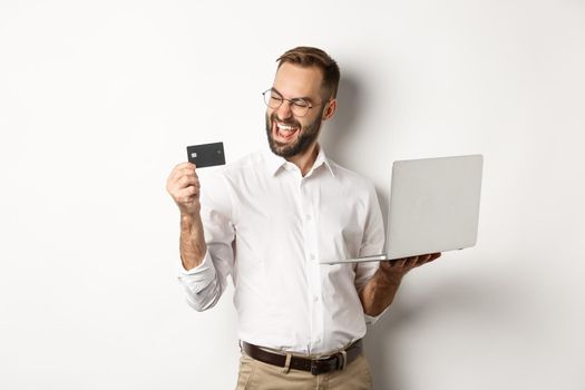 Online shopping. Satisfied handsome man looking at credit card after making order internet, using laptop, standing over white background.