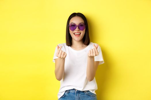 Fashion and lifestyle concept. Attractive asian woman in heart-shape sunglasses, showing finger hearts and smiling happy at camera, standing over yellow background.