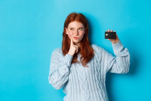 Image of thoughtful redhead girl thinking about shopping, showing credit card and pondering, standing over blue background.