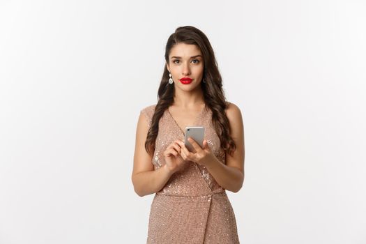 Christmas party and celebration concept. Attractive woman in luxurious dress making phone call, texting message on smartphone, standing over white background.