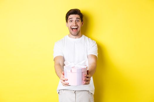 Concept of holidays and celebration. Happy man giving a gift and looking excited, standing against yellow background.