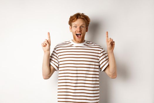 Amazed and excited redhead man checking out promotion, pointing fingers up and showing logo, staring at camera, standing over white background.