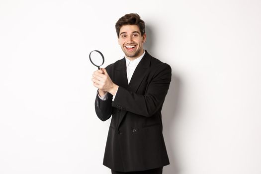 Handsome businessman in black suit, holding magnifying glass and smiling, looking for you, standing against white background.