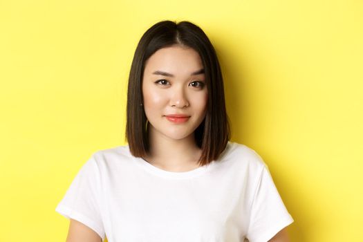 Close up of beautiful asian woman with casual makeup, raising eyebrow and looking intrigued at camera, standing curious over yellow background.
