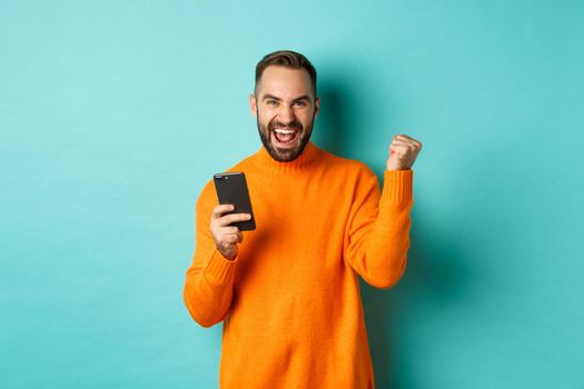 Photo of happy man triumphing, holding mobile phone and make winner gesture, rejoice and achieve goal, standing over light blue background.