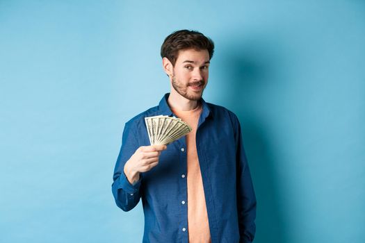 Unbothered rich guy showing cash and smiling, holding dollars in hand, standing on blue background. Copy space