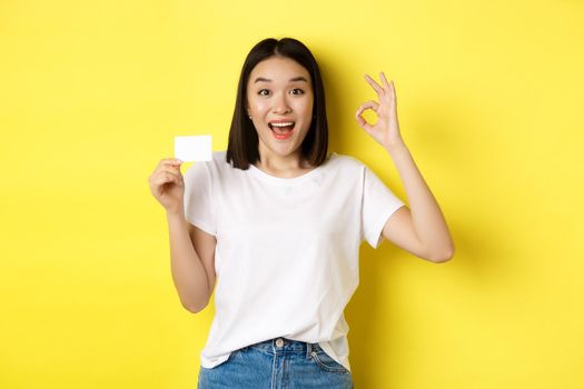 Young asian woman in casual white t-shirt showing plastic credit card and okay gesture, recommend bank, smiling at camera, yellow background.