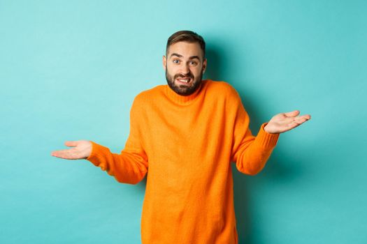 Confused caucasian man with beard, shrugging and looking clueless, standing indecisive against turquoise background.