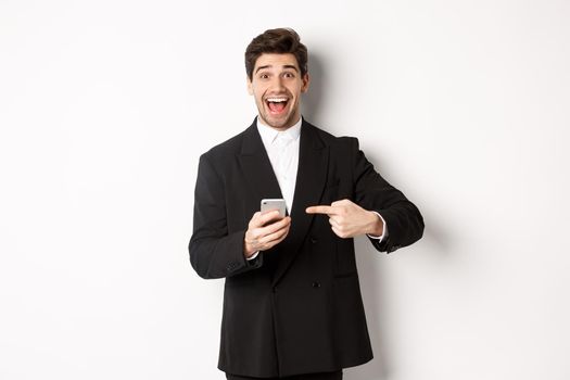 Image of cheerful businessman looking amazed, pointing at mobile phone, standing in suit over white background.