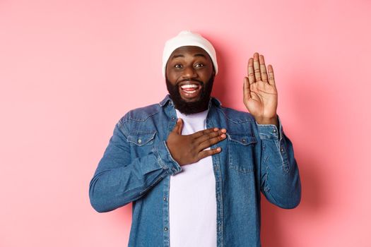 Image of smiling african-american man being honest, telling truth, hands on heart and in air, making promise or swearing, standing ove rpink background.