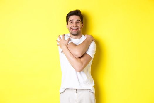 Happy kind guy hugging himself and smiling, standing pleased against yellow background. Copy space