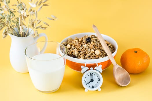 Healthy hearty breakfast. Baked granola in bowl, glass of milk, orange, wooden spoon and alarm clock on yellow background