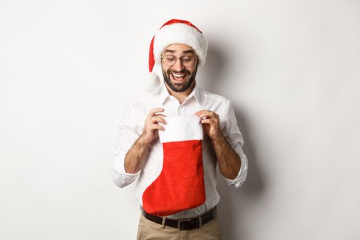 Happy adult man open christmas sock and looking inside, receiving xmas gifts for winter holidays, standing in santa hat, white background.