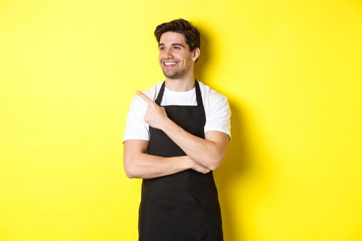 Handsome barista pointing and looking left at promo, wearing black apron, standing against yellow background.