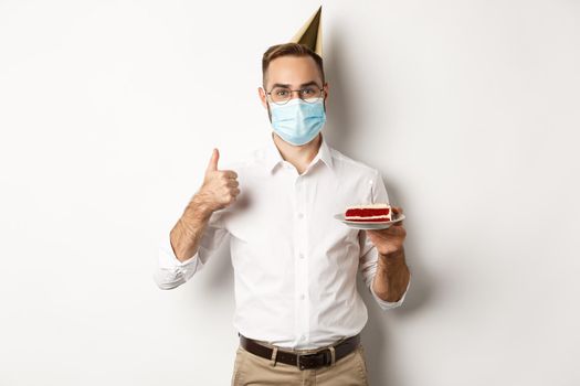 Covid-19, social distancing and celebration. Happy young man in face mask, enjoying birthday party, holding bday cake and make thumb up gesture, white background.