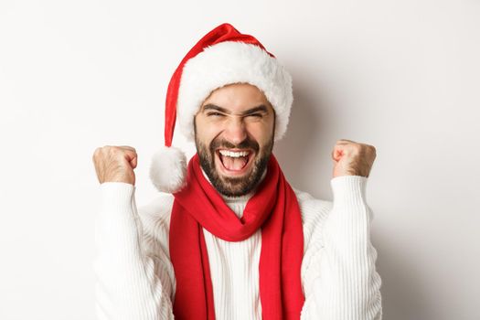 New Year party and winter holidays concept. Close-up of excited man in Santa hat rejoicing, winning or celebrating in Santa hat, standing against white background.