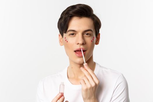 Close-up of gender fluid person with glitter on face, applying lip gloss lipstick and looking at camera, standing against white background.