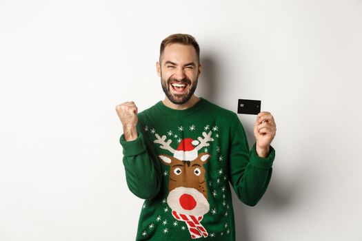 Christmas holidays and shopping concept. Excited man showing credit card and looking happy, standing over white background.