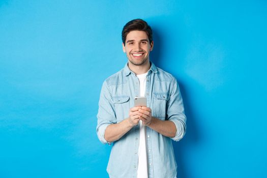 Handsome bearded man in casual outfit smiling at camera, checking smartphone, standing against blue background.