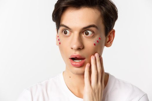 People, lgbtq and beauty concept. Headshot of gay man with glitter on face, looking left in awe, express surprise and disbelief, standing against white background.