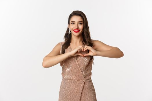 Celebration and party concept. Beautiful young woman in elegant dress showing heart sign, I love you gesture, express sympathy and smiling, white background.