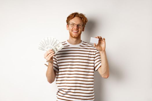 Shopping and finance concept. Young redhead man with beard and glasses showing plastic credit card with money in dollars, white background.