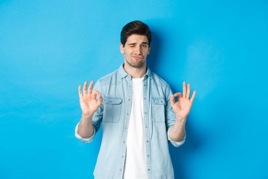 Satisfied handsome man showing a-ok signs and looking pleased, approving something good, standing against blue background.