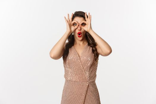 Christmas party and celebration concept. Woman in glamour dress and red lips looking at something amazing, staring through hand binoculars, standing over white background.