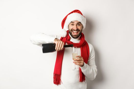 Christmas party and holidays concept. Happy bearded man in Santa hat and scard, pour himself champagne and smiling, celebrating New Year, standing over white background.