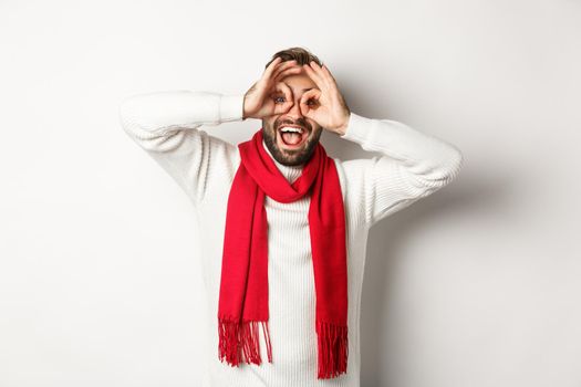 Happy guy celebrating christmas, showing funny faces and laughing, standing over white background in winter sweater and red scarf.