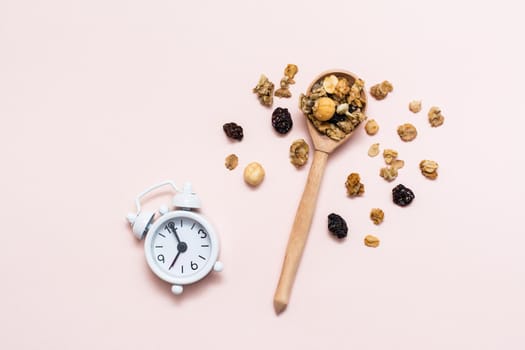 Healthy eating. Baked granola from oats, nuts and raisins in a wooden spoon and an alarm clock on a pink background. Top view