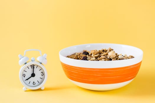 Healthy eating. Baked oats, nuts and raisins granola in bowl and alarm clock on yellow background