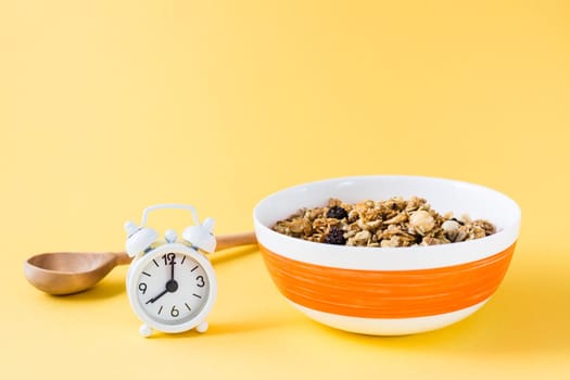 Healthy eating. Baked oats, nuts and raisins granola in bowl, wooden spoon and alarm clock on yellow background