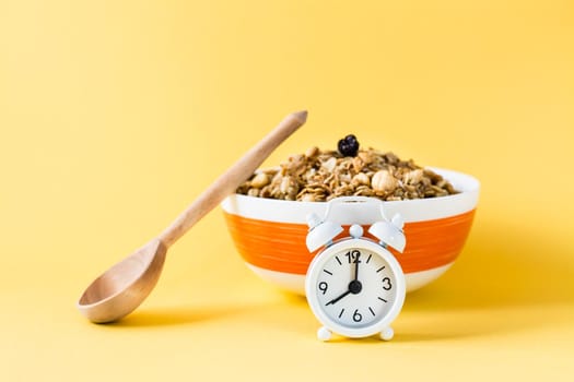 Healthy eating. Alarm clock in front of a baked granola made from oats, nuts and raisins in a bowl and a wooden spoon on a yellow background