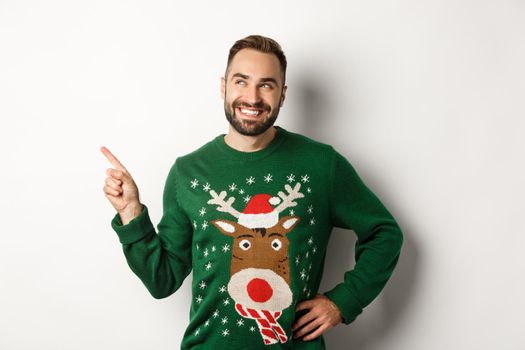New year celebration and winter holidays concept. Handsome smiling man in green christmas sweater, pointing and looking at upper left corner logo store, white background.