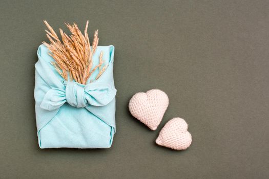 Eco friendly furoshiki gift with ears of dry grass and two knitted hearts on a green background