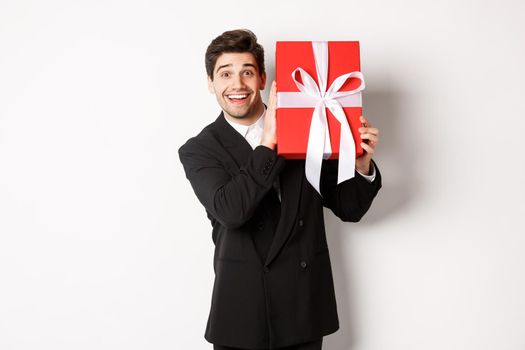 Handsome man in black suit, receiving christmas gift, smiling amazed, standing against white background.