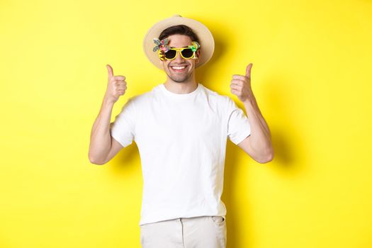 Concept of tourism and lifestyle. Image of smiling tourist showing thumbs-up, enjoying trip and recommending, wearing summer hat and sunglasses, yellow background.