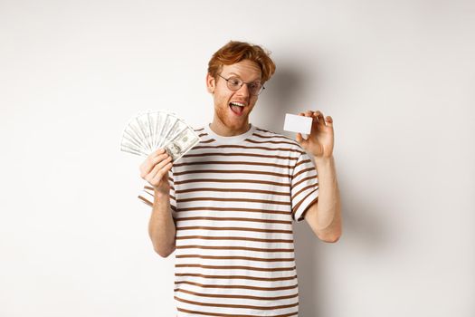 Shopping and finance concept. Young redhead man in glasses choosing plastic credit card, showing dollars and smiling, standing over white background.