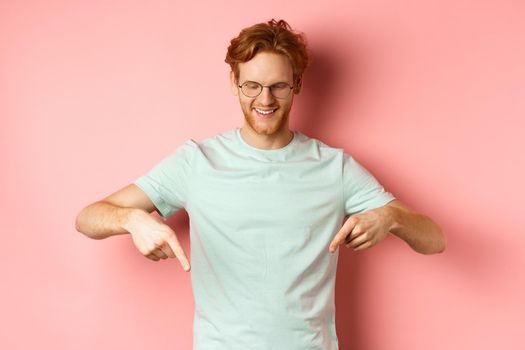 Image of handsome caucasian man with red messy hair and glasses, pointing fingers and looking down at promo offer, smiling pleased, standing over pink background.