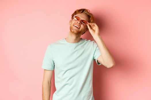 Tourism and vacation concept. Happy young man with red hair relaxing, wearing sunglasses and smiling carefree, standing over pink background.